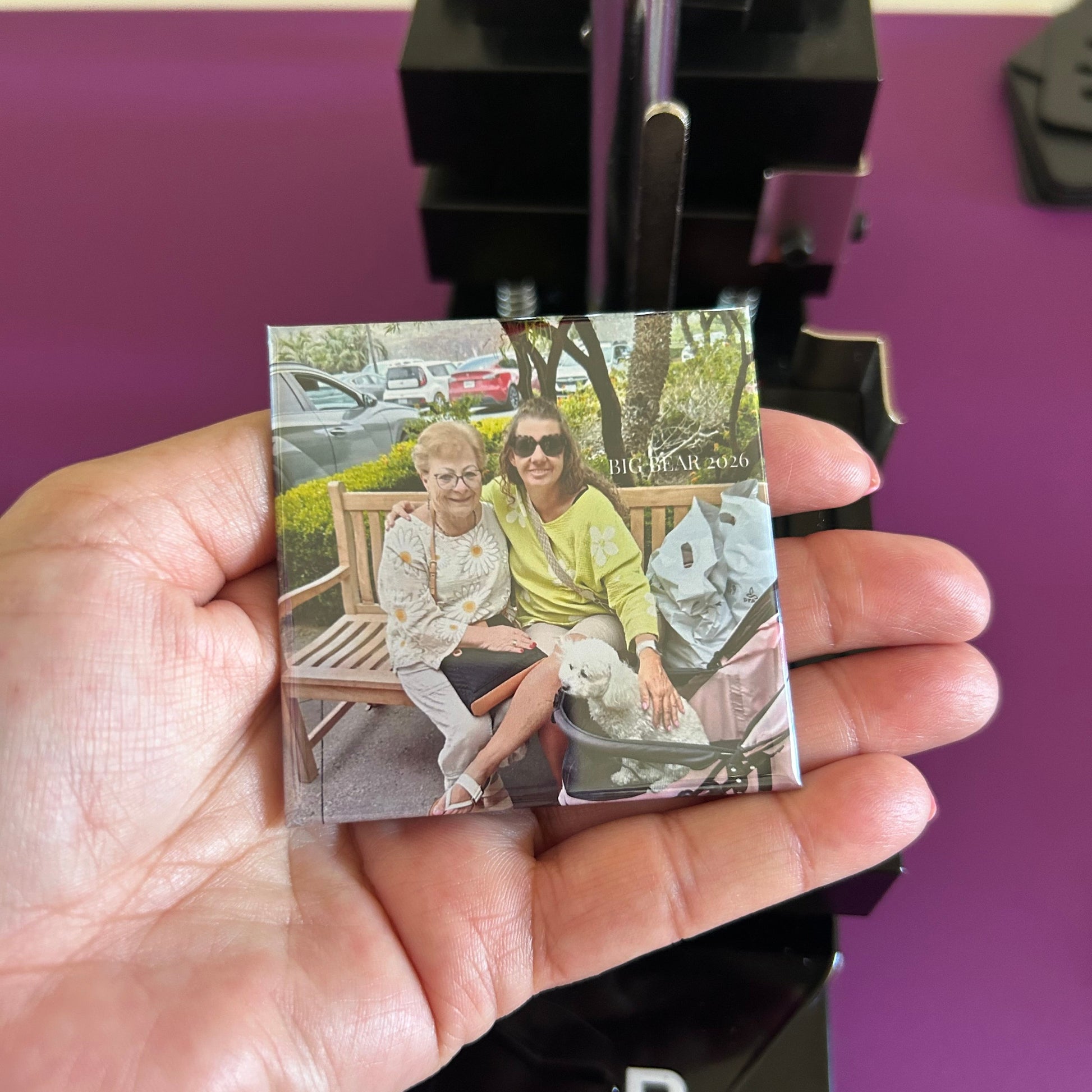 Hand holding a square fridge magnet with a colorful photo of two people sitting on a bench.