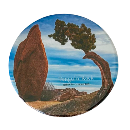 Square coaster featuring a photo of Penguin Rock at Joshua Tree National Park — a tall pointed boulder beside a wind-bent juniper tree under a vivid blue sky.