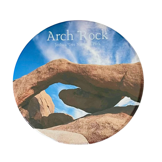 Square coaster featuring a close-up photo of Arch Rock at Joshua Tree National Park — a sweeping granite arch framing a bright blue sky with wispy clouds.