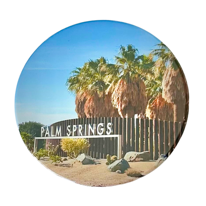 Round 3.5" coaster featuring the iconic Palm Springs sign with bold white lettering, towering palm trees, desert landscaping, and a bright blue California sky.