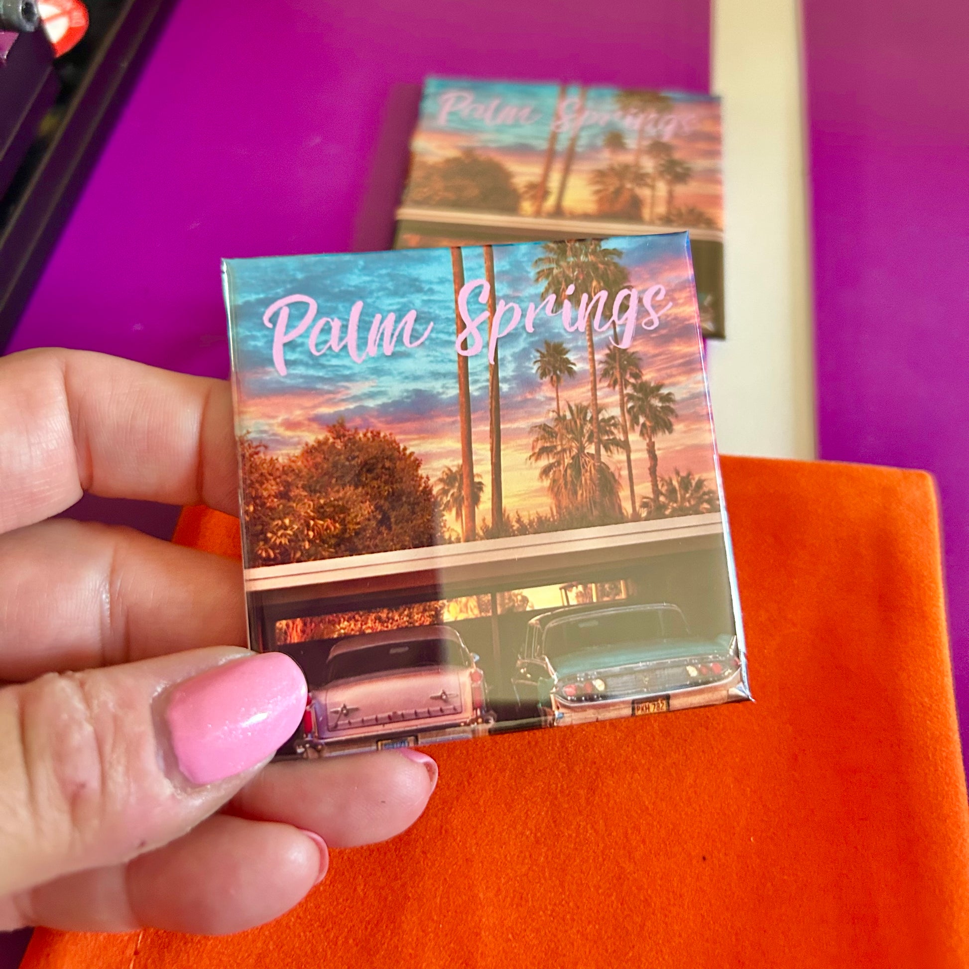 Hand holding a 'Palm Springs' themed magnet with palm trees and vintage cars on a vibrant background.